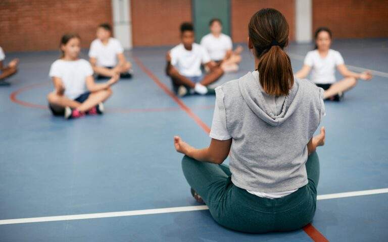 Vista trasera de una profesora de deportes practicando Yoga con sus alumnos en el gimnasio de la escuela.